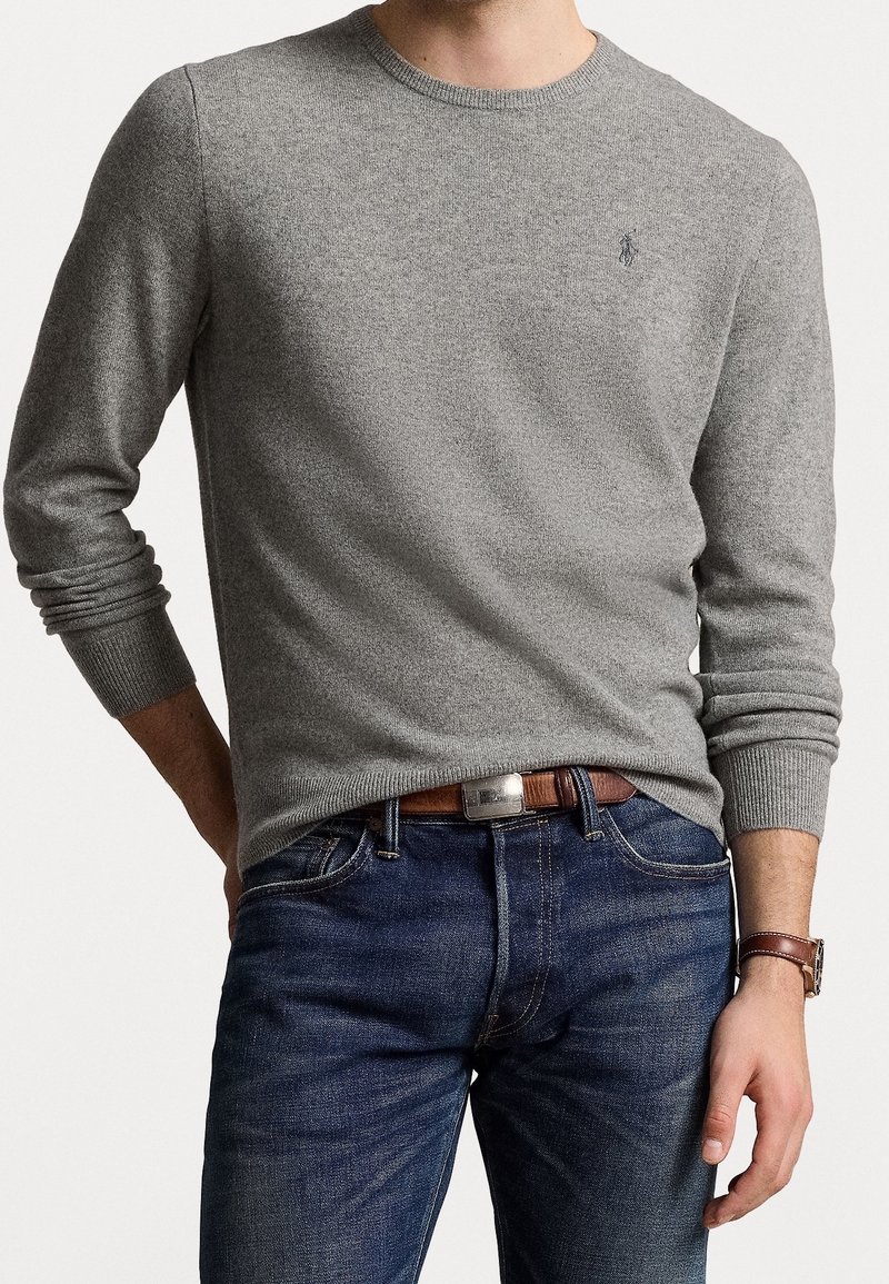 Pull gris à col rond en tissu texturé, avec manches longues et petit logo sur la poitrine, associé à un jean denim foncé et une ceinture marron.