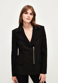 Schwarzer Blazer mit scharfem Revers, silbernem Reißverschluss, zwei vorderen Taschen und tailliertem Schnitt. Glattes, strukturiertes Material.