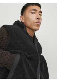 Schwarze Fleece-Weste mit strukturierter Oberfläche und hohem Kragen. Mit kontrastierenden schwarzen Reißverschlüssen und einem gemütlichen braunen Strickpullover darunter.