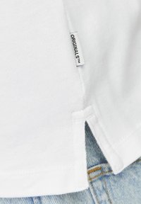 Tricou alb din bumbac, cu o fanta pe lateral si o eticheta inscriptionata "ORIGINALS™." Textura materialului este fina, cu un design usor.