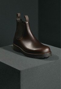 Dunkelbrauner Chelsea-Stiefel aus Leder mit elastischen Seitenteilen und Zuglaschen, präsentiert auf einem grauen Podest vor dunklem Hintergrund.