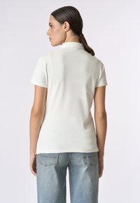 Polo shirt bianco con maniche corte, dotato di colletto e una forma aderente. La texture è liscia, abbinata a jeans azzurri chiari e morbidi.