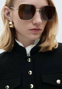 Brown-tinted oversized sunglasses with a gold metal frame; model has shoulder-length blonde hair and wears a black jacket with gold buttons.