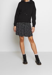 Sweatshirt à capuche noir avec cordons, assorti à une jupe noire à pois. Le mannequin porte des bottines noires à lacets et des chaussettes blanches à la cheville.