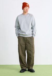 Sweat-shirt gris avec des poignets côtelés, pantalon cargo olive avec poches latérales et chaussures noires. Un bonnet orange avec un logo complète la tenue.