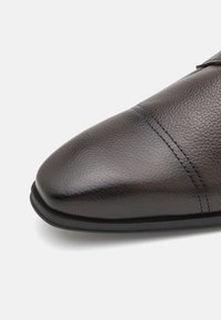 Chaussure en cuir marron avec une surface texturée, un bout pointu et un détail de couture subtil. Semelle en caoutchouc noire au profil élégant.