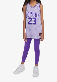 Canotta da basket in tessuto rete viola con "JORDAN" e "23" in viola scuro, abbinata a leggings viola e sneakers bianche.