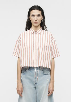 Kobieta o długich ciemnych włosach, nosząca złote kolczyki-koła, białą koszulę z krótkim rękawem i pionowymi czerwonymi paskami oraz jasnoniebieskie podarte dżinsy.