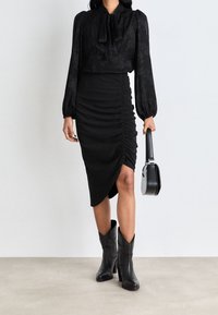 Schwarze Bluse mit gebundenem Ausschnitt und floralem Muster, kombiniert mit einem gerafften schwarzen Rock mit seitlichem Schlitz. Getragen mit schwarzen Stiefeln mit Absatz.