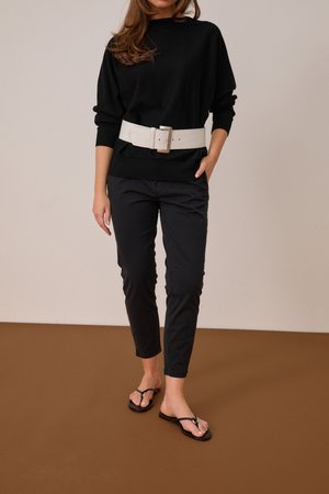 Kvinde iført sort sweater, sorte bukser, bredt hvidt bælte og sorte sandaler, stående mod en ensfarvet baggrund.