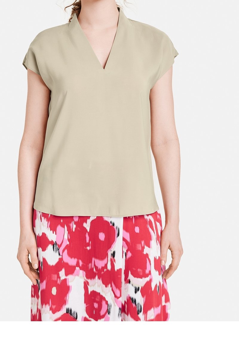 Kortärmad blus i ljus khaki med V-ringning, tillverkad av ett slätt tyg; bärs med en livligt mönstrad rosa och vit kjol.