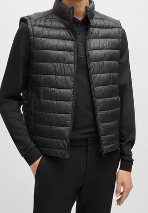 Mann trägt eine schwarze gesteppte Weste mit Reißverschluss über einem schwarzen Polo mit langen Ärmeln, mit einer Hand in der Tasche vor einfachem Hintergrund.