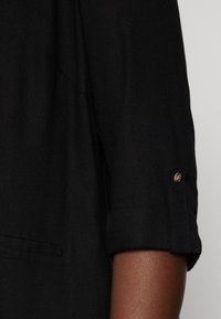 Camicia in tessuto nero con maniche arrotolate, caratterizzata da un dettaglio a bottoni. Texture liscia e vestibilità su misura con tasca frontale.