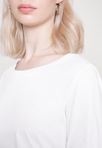 Weißes Baumwoll-T-Shirt mit weitem Ausschnitt. Kurze Ärmel zeigen einen glatten Halsausschnitt. Ergänzt durch goldene Creolen mit dekorativen Akzenten.