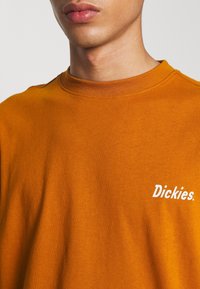 Orange cotton sweatshirt with a round neckline and a white "Dickies." logo on the left chest. Smooth texture and casual fit.