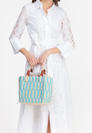 Tkaná taška přes rameno s modro-beige pruhy, provazovými uchy a dřevěnými akcenty. Taška má stahovací uzávěr a kompaktní čtvercový tvar.