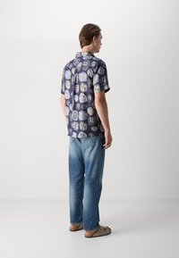 Camisa de manga curta, azul marinho, com um padrão de círculos e formas, combinada com jeans azul claro e sandálias castanhas, vista pela parte de trás.