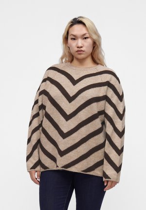 Personne aux longs cheveux blonds portant un pull beige avec des rayures chevrons brun foncé et un pantalon sombre, se tenant devant un fond uni.