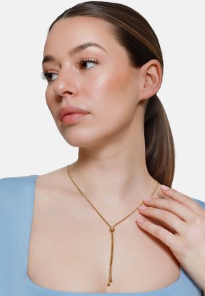 Frau mit glatter Haut, die ein hellblaues Oberteil trägt und eine zarte goldene Lariat-Kette mit einem gleitenden Perlen- und Quasten-Anhänger anlegt.