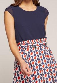 Blouse bleu marine à manches courtes et transparentes associée à une jupe à motifs avec des formes en zigzag rouges, blanches et bleues, accompagnée d'une ceinture.