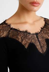 Haut noir avec un décolleté festonné orné de dentelle noire intricte. Texture douce avec des détails de couture visibles autour de la zone du col.