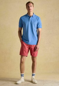 Sinine puuvillane polo särk klassikalise kraega, koos roosade lühikeste pükstega. Mudel kannab valgeid spordijalatsite ja triibulisi sokke. Lihtne, igapäevane disain.