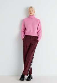 Pink knitted turtleneck sweater paired with maroon trousers featuring side stripes. Shoes are black, thick-soled, and textured.