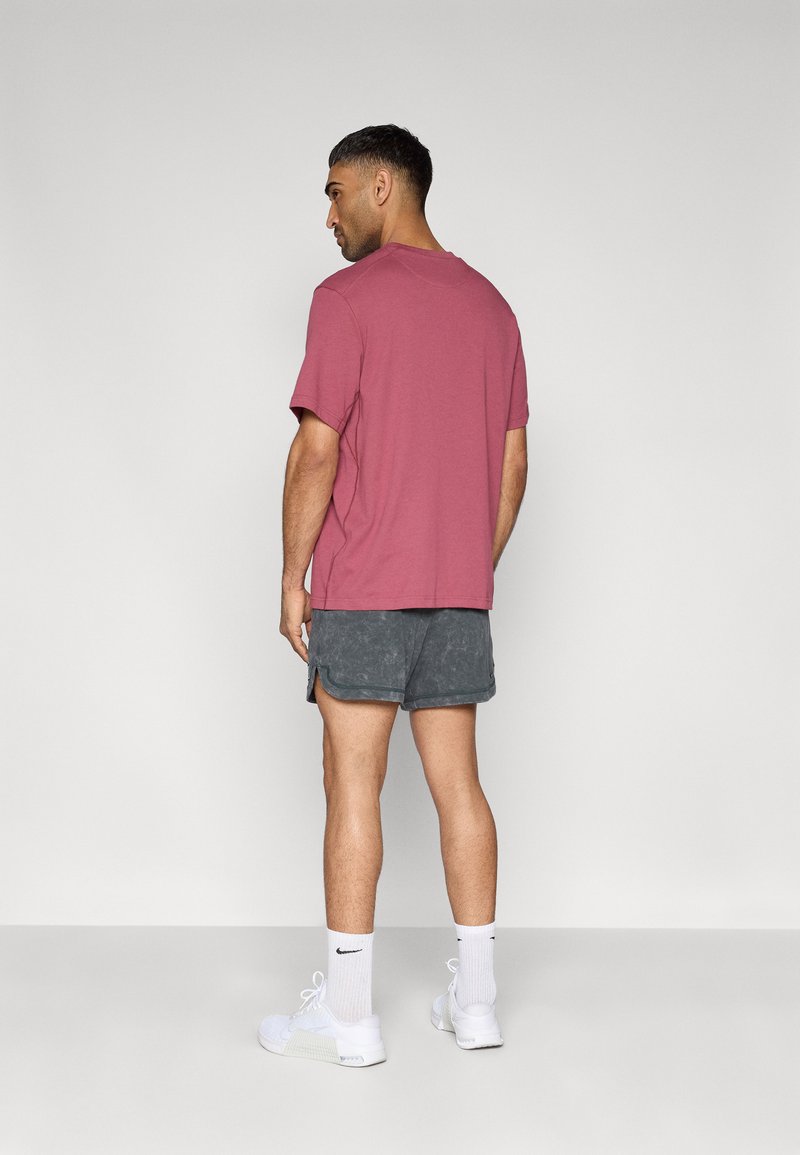 Bordeauxrotes Baumwoll-T-Shirt, kurzärmelig, locker geschnitten; kombiniert mit dunkelgrauen, strukturierten Shorts und weißen Sneakers mit schwarzen Akzenten.