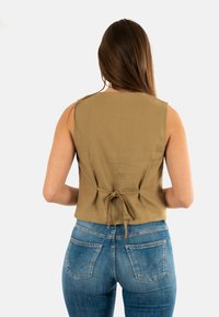 Top sin mangas color caqui con un detalle de lazo en la parte trasera, combinado con unos jeans de mezclilla azul que presentan un diseño de bolsillo distintivo. Textura de tela suave.