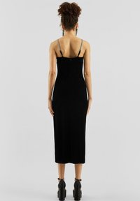 Žena s kovrčavom kosom u visokom punđu nosi usku crnu midi haljinu s tankim naramenicama i crne platforme, okrenuta leđima.