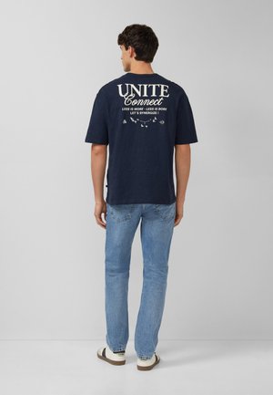 Homme portant un t-shirt bleu marine avec le texte « UNITE Connect », un jean bleu clair et des baskets blanches, debout devant un fond gris uni.