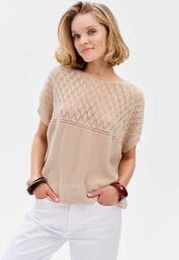 Beżowy sweter z dzianiny z diamentowym wzorem na górze, gładka faktura na dole, krótkie rękawy, zestawiony z białymi spodniami i bransoletkami.