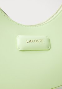 Zelena gumena torba s istaknutim zlatnim logotipom "LACOSTE" na teksturiranoj, mat površini, s zatvaračem duž gornje ivice.