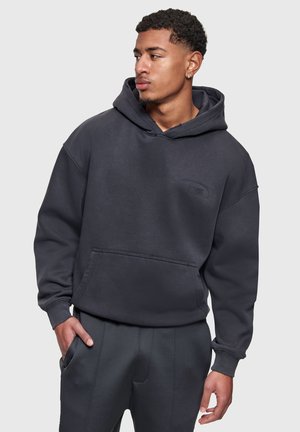 Jeune homme portant un sweat à capuche noir avec une poche avant et un pantalon sombre, regardant vers la gauche avec une main dans sa poche.