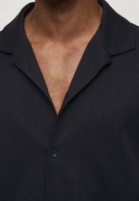 Primo piano di una persona che indossa una camicia nera con colletto e il primo bottone slacciato, che rivela parte del loro petto superiore e del collo.