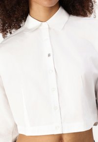 Camicia bianca corta realizzata in tessuto liscio, caratterizzata da un colletto classico, chiusura con bottoni e un piccolo logo vicino ai bottoni.