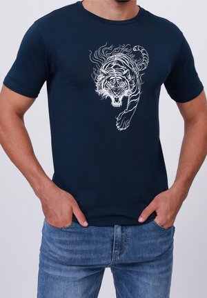 Mann trägt ein dunkelblaues T-Shirt mit weißem Grafikdruck eines brüllenden Tigers in Bewegung auf der Vorderseite, kombiniert mit blauen Jeans, Hände in den Taschen.