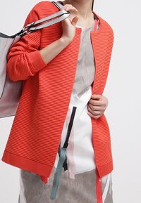 Röd ribbad cardigan med öppen front, parat med en mönstrad grå klänning med färgade detaljer. Modellen håller en silverfärgad handväska.