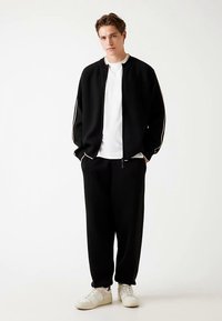 Veste noire à fermeture éclair avec bordure blanche, associée à un pantalon noir et un t-shirt blanc. Le modèle porte des baskets blanches et pose devant un fond neutre.