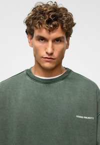 Sweat-shirt à col ras du cou vert olive en tissu doux, avec un col côtelé et un petit logo blanc sur la partie gauche de la poitrine.