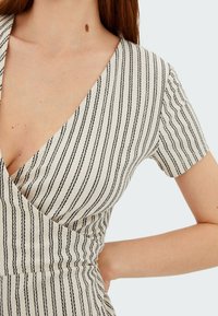 Femme portant une robe beige avec des rayures verticales noires, à manches courtes et décolleté profond en V, la main posée sur la taille.