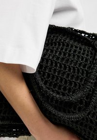 Schwarze geflochtene Handtasche mit strukturiertem Design, runden Akzenten und einer weichen, strukturierten Form, die unter einem weißen Ärmeln gehalten wird.