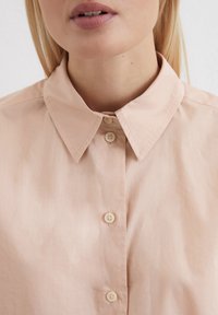 Helles rosa Bluse mit spitzem Kragen, mit einer Knopfleiste vorne und zwei sichtbaren Knöpfen. Glatte Textur und minimales Design.
