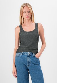 Graues, ärmelloses Oberteil mit quadrischem Ausschnitt, aus einem strukturierten Stoff gefertigt. Kombiniert mit hochgeschlossenen blauen Jeans.