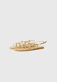Gold sandals with intricate crisscross straps and a flat sole. Features adjustable ankle straps and a smooth, shiny finish.