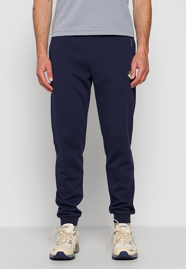 Granatowe dresy wykonane z miękkiego materiału, z elastycznym paskiem w talii, mankietami oraz małym zielonym logo na boku.