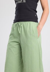 Pantalon de pyjama à carreaux verts et blancs avec une taille élastique et un cordon de serrage, doté de poches latérales et d'un design à jambes larges.