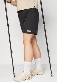 Black shorts with a silver logo, synthetic material, casual fit, paired with light-colored hiking shoes and trekking poles.
