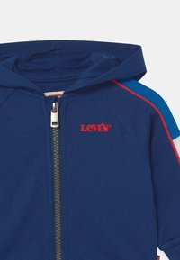 Chaqueta con capucha azul y logo rojo de Levi's, con cremallera en la parte frontal y franjas contrastantes en azul, rojo y blanco a lo largo de los hombros.