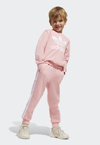 Ružová mikina Adidas s bielym logom, skombinovaná s rovnakými ružovými joggermi s bielymi pruhmi na bokoch. Textúrovaná tkanina a voľný strih.